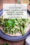 A plate with flatbread topped with a green salad mixture, tender chunks of salmon, and a large basil leaf, garnished to perfection—this chopped green goddess salmon flatbread is described as macro-friendly and irresistibly fresh.