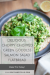 A chopped Green Goddess salmon flatbread is topped with vibrant green salad, tender salmon, and fresh basil, all arranged on a dark plate. A lemon half and greens are in the background. Text overlay promotes a Green Goddess salmon salad flatbread recipe.