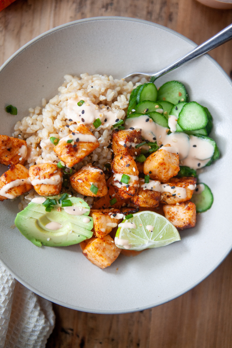 Gray bowl filled with brown rice, sliced cucumbers, green chives, sriracha mayo, sliced avocado, sliced lime, and air fryer salmon bites.