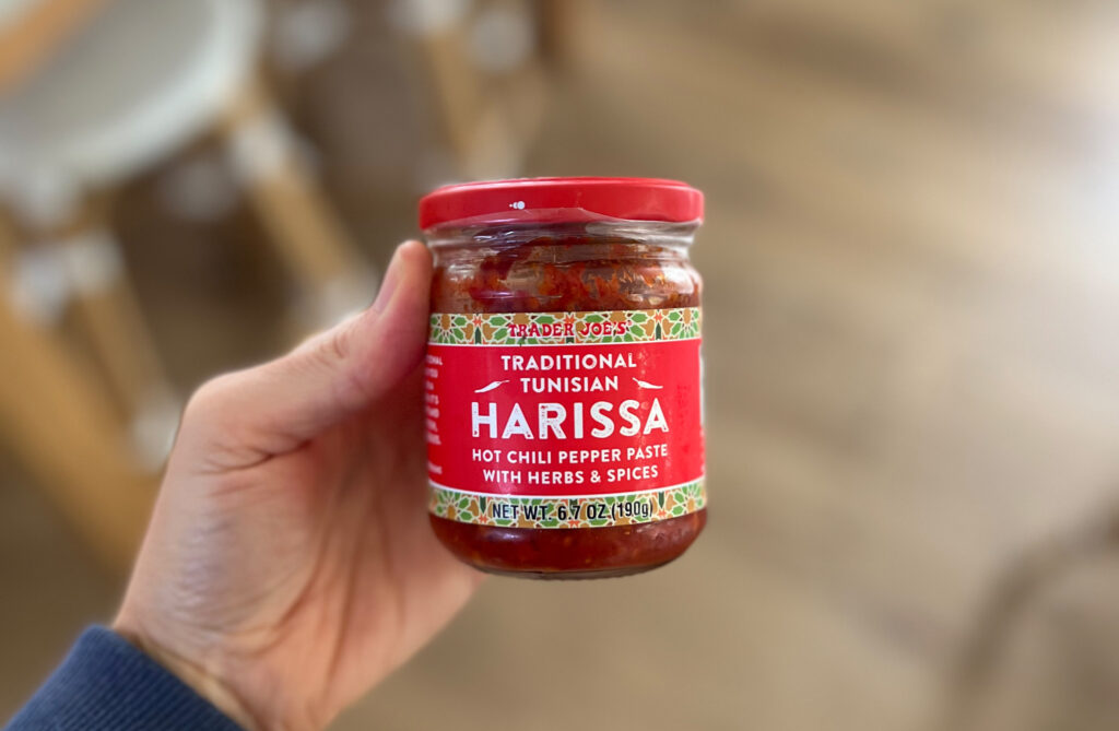 A hand holds a jar of Trader Joe’s Traditional Tunisian Harissa hot chili pepper paste with herbs and spices. The label is red with white and yellow text, making it one of My Favorite Trader Joe’s Products for adding bold flavor to any dish.