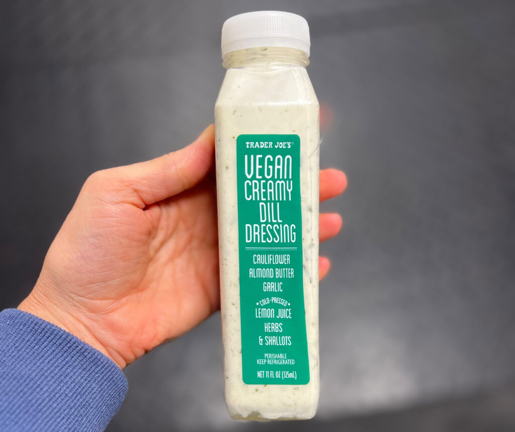 A hand holds a bottle of Trader Joe’s Vegan Creamy Dill Dressing, with a green label listing ingredients like cauliflower, almond butter, garlic, lemon juice, herbs, and shallots—one of My Favorite Trader Joe’s Products.