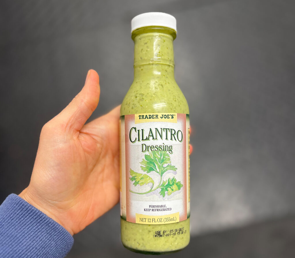 A hand holds a bottle of Trader Joe’s Cilantro Dressing against a blurred dark background. The bottle is green with a white cap and label featuring cilantro leaves and product details, making it one of My Favorite Trader Joe’s Products.