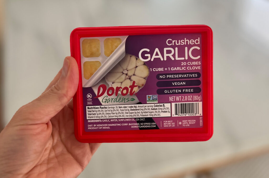 A hand holding a red container of Dorot Gardens Crushed Garlic—one of My Favorite Trader Joe’s Products—shows 20 cubes with labels for vegan, gluten free, no preservatives, and each cube equals one garlic clove.