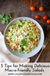 A bowl of chopped salad with tomatoes, greens, and cheese sits on a wooden table with a fork, napkin, herbs, and cherry tomatoes nearby. Text reads: 5 Secrets to Amazing Macro-friendly Salads—discover how to make your macro-friendly salads both nutritious and delicious.