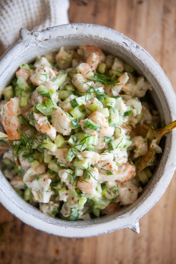 A bowl of cold shrimp salad with chopped celery, cucumbers, and fresh dill in a creamy dressing, placed on a wooden table.