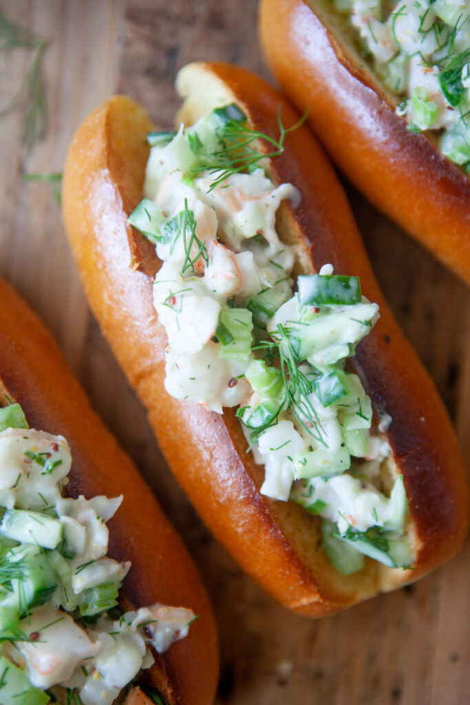 Three toasted buns filled with a creamy cold shrimp salad made of chopped vegetables and herbs, garnished with fresh dill, arranged on a wooden surface.