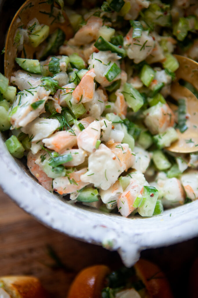 A close-up of a creamy cold shrimp salad in a bowl, featuring succulent shrimp, diced vegetables, fresh herbs, and a light dressing, with a wooden spoon partially visible.