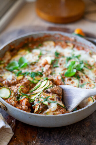 A skillet filled with Skillet Zucchini Lasagna, topped with melted cheese and fresh herbs, sits on a wooden table. A wooden spoon lifts a serving, revealing delicious layers of zucchini and savory sauce.