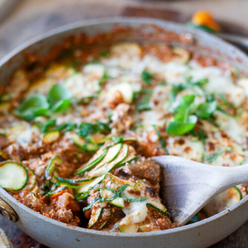 A skillet filled with Skillet Zucchini Lasagna, topped with melted cheese and fresh herbs, sits on a wooden table. A wooden spoon lifts a serving, revealing delicious layers of zucchini and savory sauce.