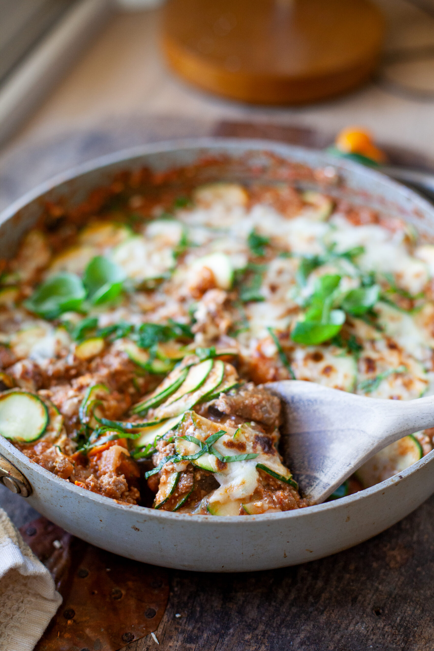 A skillet filled with Skillet Zucchini Lasagna, topped with melted cheese and fresh herbs, sits on a wooden table. A wooden spoon lifts a serving, revealing delicious layers of zucchini and savory sauce.