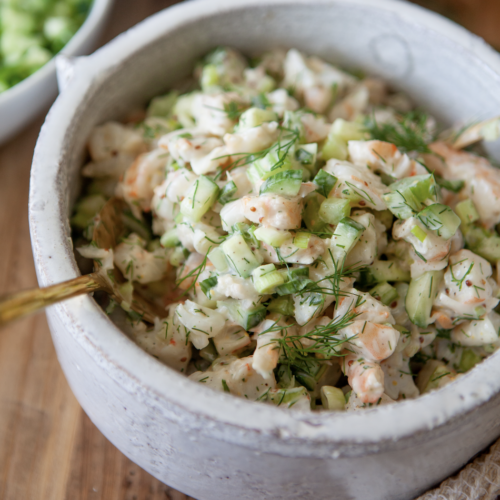 A ceramic bowl filled with Cold Shrimp Salad featuring chopped celery, fresh dill, and a creamy dressing, with a gold spoon inside.