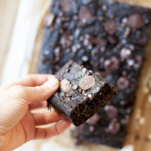 A hand holds a piece of chocolate pumpkin snack cake above a tray of brownies topped with chocolate chips. The brownies rest on parchment paper.