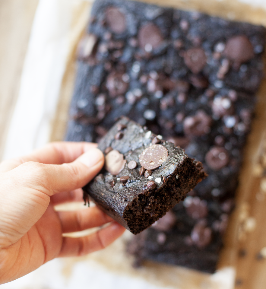A hand holds a piece of chocolate pumpkin snack cake above a tray of brownies topped with chocolate chips. The brownies rest on parchment paper.