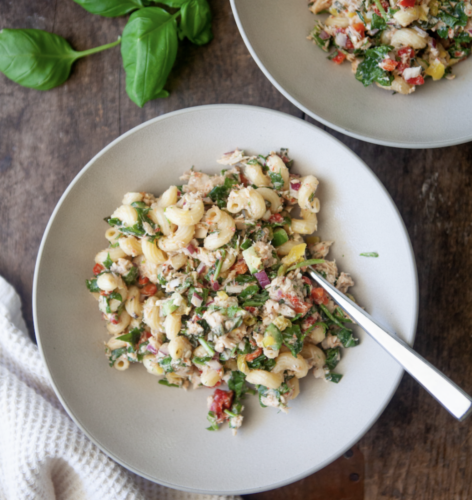A vibrant bowl of tuna pasta salad with spinach, red and yellow peppers, and fresh herbs sits on a wooden table with a fork. Fresh basil leaves and another bowl of salad are arranged nearby.