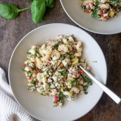 A vibrant bowl of tuna pasta salad with spinach, red and yellow peppers, and fresh herbs sits on a wooden table with a fork. Fresh basil leaves and another bowl of salad are arranged nearby.