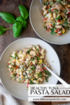 Two bowls of tuna pasta salad with spinach and peppers sit on a wooden table, garnished with fresh basil. Text on the image reads Healthy Tuna Pasta Salad www.lillieeatsandtells.com.