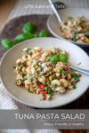 A bowl of tuna pasta salad brimming with colorful vegetables and fresh herbs sits invitingly on a table. In the background, a second bowl of this vibrant tuna pasta salad is softly blurred, adding depth to the scene. Text on the image reads Tuna Pasta Salad, Macro-Friendly & Healthy.