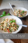 Two bowls of tuna pasta salad with herbs sit on a table, a fork resting in one bowl. A text overlay reads Tuna Pasta Salad that’s macro-friendly & healthy, highlighting the balanced nutrition of this wholesome dish. Fresh basil leaves are placed beside the bowls, adding a vibrant touch to the inviting tuna pasta salad spread.