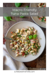 A bowl of tuna pasta salad with vegetables and herbs, held by a hand with a fork. Another bowl of the vibrant tuna pasta salad sits nearby on a wooden table, garnished with fresh basil. Text reads: Macro-Friendly Tuna Pasta Salad.