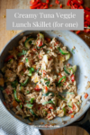 A skillet filled with a creamy tuna and vegetable mixture, including zucchini and red pepper, sits on a wooden surface. Chopped red pepper is scattered nearby. Text above reads: Creamy Tuna Veggie Lunch Skillet for One, highlighting this easy and flavorful single-serving meal.
