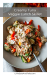 Bowl of creamy tuna and mixed vegetables with a fork, topped with grated cheese, on a wooden table. Text overlay reads Creamy Tuna Veggie Lunch Skillet for One.