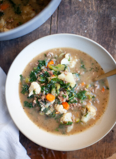 A bowl of Macro-Friendly Beef and Kale Soup with ground meat, cauliflower, carrots, kale, and broth sits on a wooden table, with a pot of soup partly visible in the background.