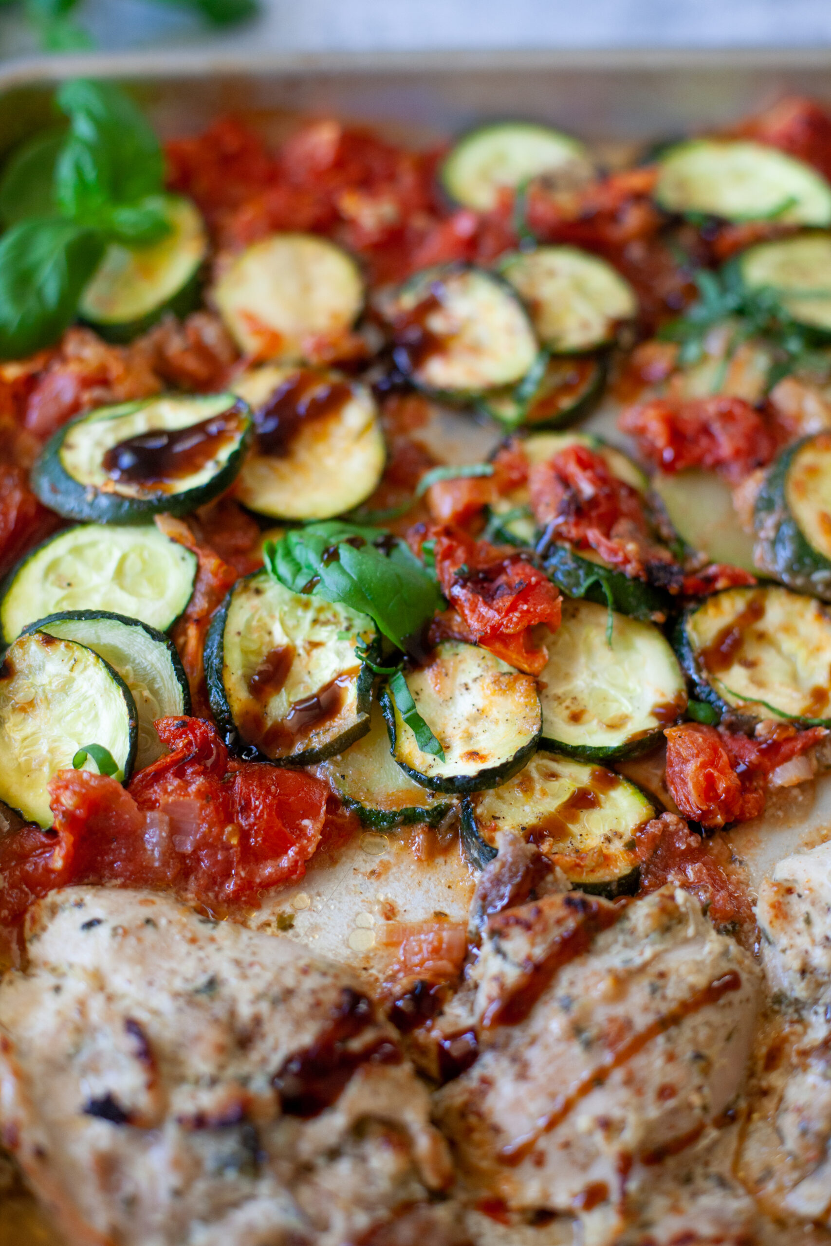 Sheet pan with Roasted Tomato Basil Chicken features seasoned chicken breasts, roasted zucchini slices, and tomatoes, all garnished with fresh basil leaves. The vegetables appear tender and slightly caramelized.