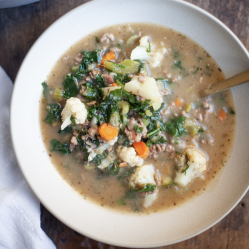 A bowl of hearty Macro-Friendly Beef and Kale Soup with ground meat, carrots, cauliflower, kale, and broth, served with a spoon on a wooden table.