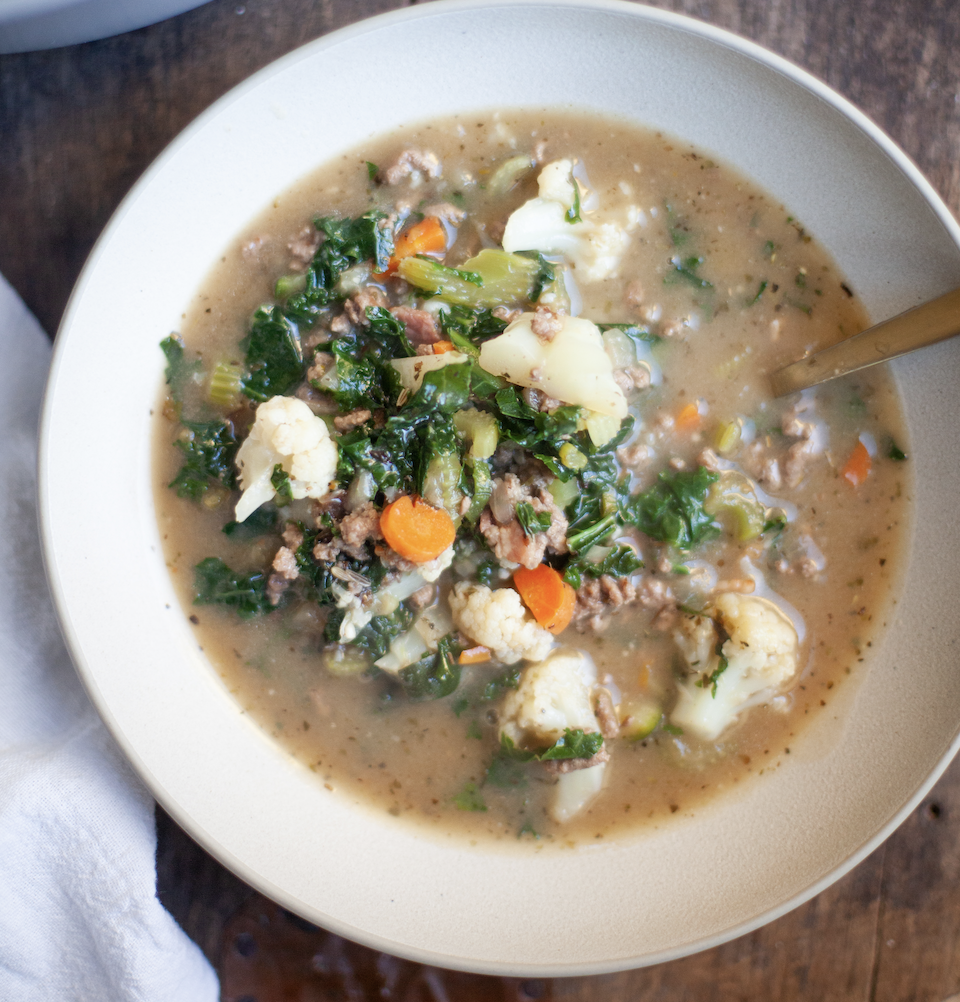 A bowl of hearty Macro-Friendly Beef and Kale Soup with ground meat, carrots, cauliflower, kale, and broth, served with a spoon on a wooden table.