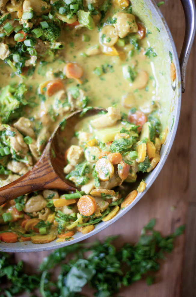 A closeup of a skillet filled with creamy, macro-friendly chicken and vegetable curry, including carrots, broccoli, and herbs, with a wooden spoon resting inside the pan. Fresh chopped herbs are scattered on the wooden surface nearby, making this an ideal choice for satisfying one-dish meals.