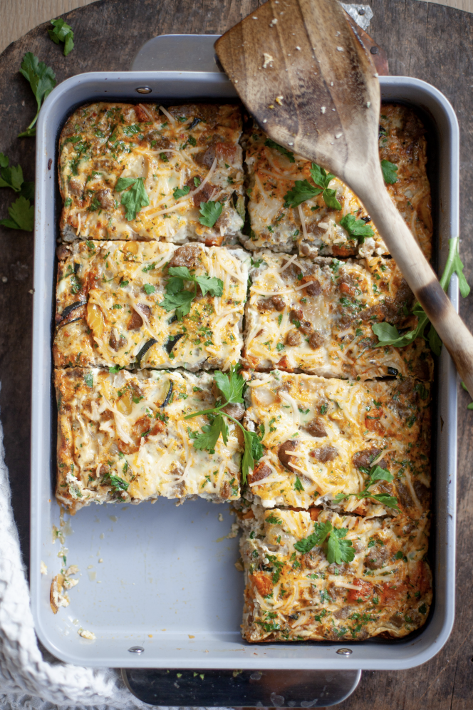 A baking dish with six pieces of a baked egg casserole topped with herbs and cheese, with one piece missing. Easy to prepare and perfect for macro-friendly one-dish meals, this wholesome breakfast or brunch option is served straight from the oven, while a wooden spatula rests on top of the casserole.