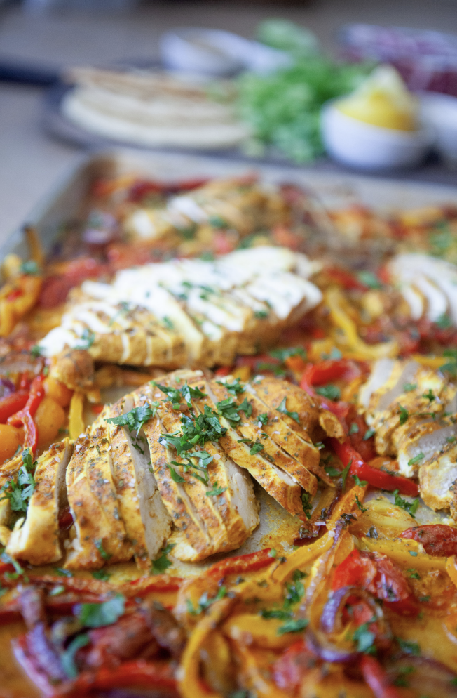 Sheet pan with sliced, seasoned chicken breast, red and yellow bell peppers, and chopped herbs creates a colorful display perfect for macro-friendly one-dish meals. In the background, pita bread, lemon wedges, and fresh greens are slightly out of focus.