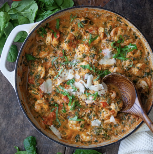 A skillet filled with creamy tomato shrimp and spinach in a luscious red pepper cauliflower cream sauce, topped with shaved parmesan and herbs, sits on a rustic wooden surface. A wooden spoon rests in the pan, and fresh spinach leaves are nearby.