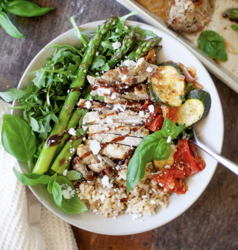 A bowl featuring Roasted Tomato Basil Chicken, asparagus, arugula, zucchini, brown rice, feta cheese, and fresh basil, all drizzled with balsamic glaze.