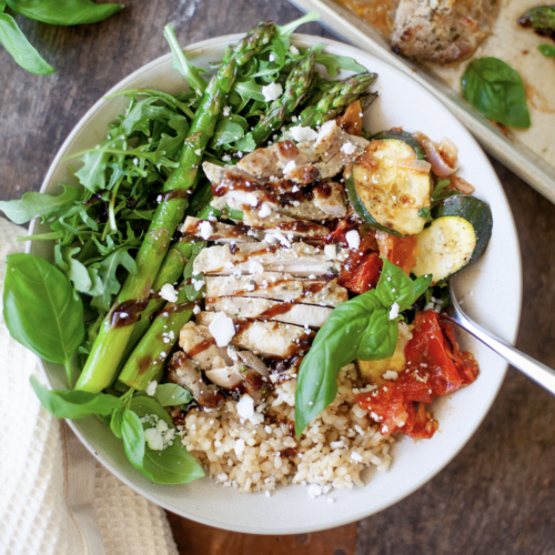 A bowl featuring Roasted Tomato Basil Chicken, asparagus, arugula, zucchini, brown rice, feta cheese, and fresh basil, all drizzled with balsamic glaze.
