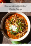 A bowl of Italian Pasta Soup with ground meat, pasta, green herbs, and a dollop of cream is shown from above on a wooden table. Text above reads Macro-friendly Italian Pasta Soup.