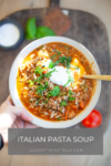 A bowl of Italian Pasta Soup topped with fresh herbs and a dollop of cream, held above a wooden board adorned with basil and tomatoes. Text at the bottom reads ITALIAN PASTA SOUP and lillieeatsandtells.com.