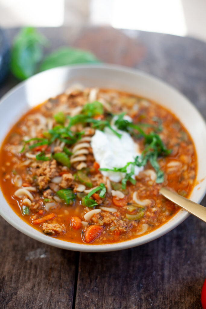 A bowl of hearty Italian Pasta Soup, featuring a rich tomato-based broth with tender pasta, savory ground meat, chopped herbs, and a dollop of sour cream, served on a rustic wooden table.