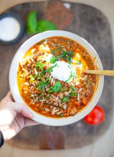 A hand holds a bowl of hearty Italian Pasta Soup with ground meat, orzo, chopped herbs, and a dollop of sour cream on top. A spoon rests in the bowl. The background includes a tomato and a small bowl.