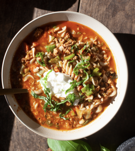 Bowl of Italian Pasta Soup with noodles, ground meat, tomato broth, topped with sour cream, chopped green onions, and fresh herbs, sitting on a wooden table in sunlight.