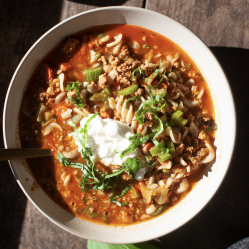 Bowl of Italian Pasta Soup with noodles, ground meat, tomato broth, topped with sour cream, chopped green onions, and fresh herbs, sitting on a wooden table in sunlight.