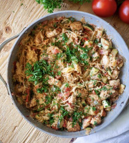 A skillet filled with a colorful Cajun Chicken Spaghetti Squash Skillet, garnished with chopped fresh parsley. Tomatoes and herbs are nearby on a rustic wooden table.