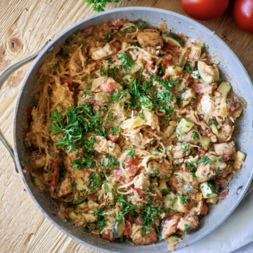 A skillet filled with a colorful Cajun Chicken Spaghetti Squash Skillet, garnished with chopped fresh parsley. Tomatoes and herbs are nearby on a rustic wooden table.