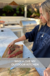 A woman seasons raw meat with herbs on a wooden board in an outdoor kitchen, highlighting why I love my outdoor kitchen. Grills and plants are visible in the background, creating a warm, inviting space. Text reads: WHY I LOVE MY OUTDOOR KITCHEN.