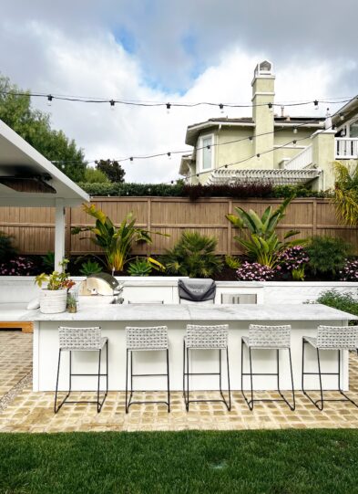 Modern outdoor kitchen with a white counter, five woven barstools, plants, string lights, and a house with a balcony in the background under a cloudy sky—just one of the reasons Why I Love My Outdoor Kitchen.