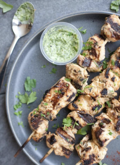 Grilled chicken skewers on a gray plate, garnished with chopped herbs, with a small container of green sauce and a spoon on the side—perfect for anyone seeking delicious options for macro-focused meal planning.