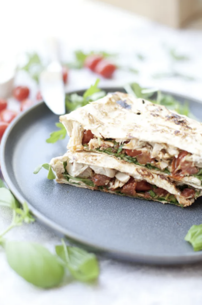 Two triangular slices of Macro-friendly Crunch Wraps, packed with greens and tomatoes, sit atop a dark plate. Fresh herbs and cherry tomatoes are scattered around the plate, adding a burst of color to this veggie-filled flatbread sandwich.