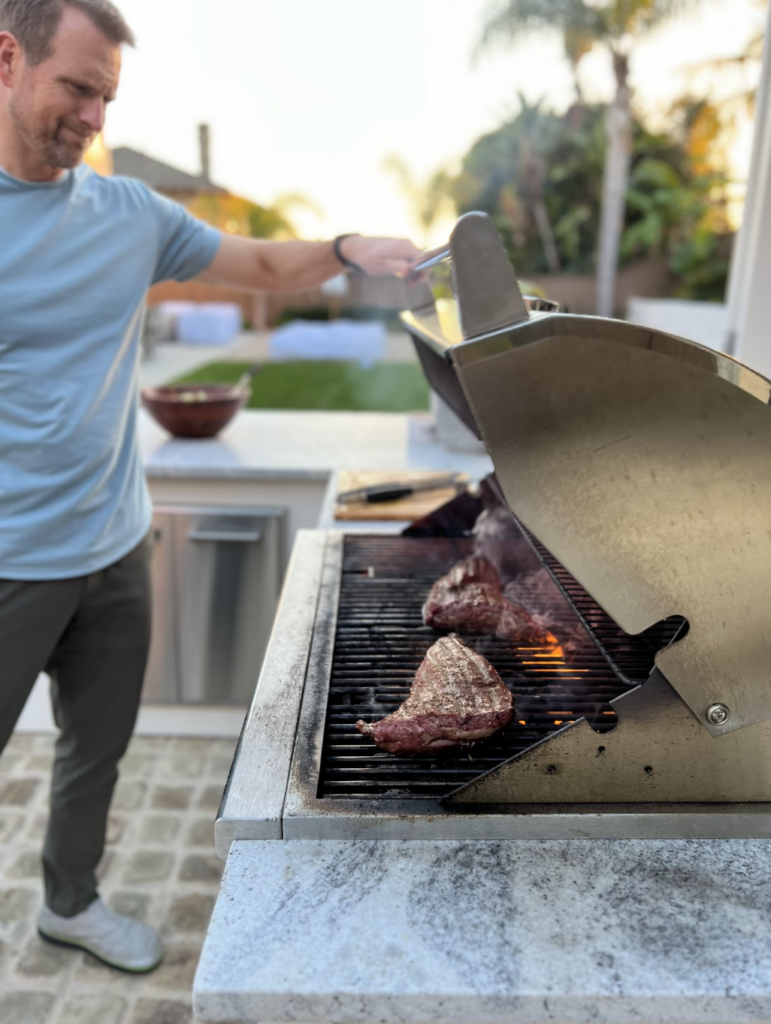 A man grills several pieces of steak on an outdoor barbecue, with flames and smoke rising from the grill. There is a bowl on a counter in the background and lush greenery in the yard—a perfect scene that captures why I love my outdoor kitchen.