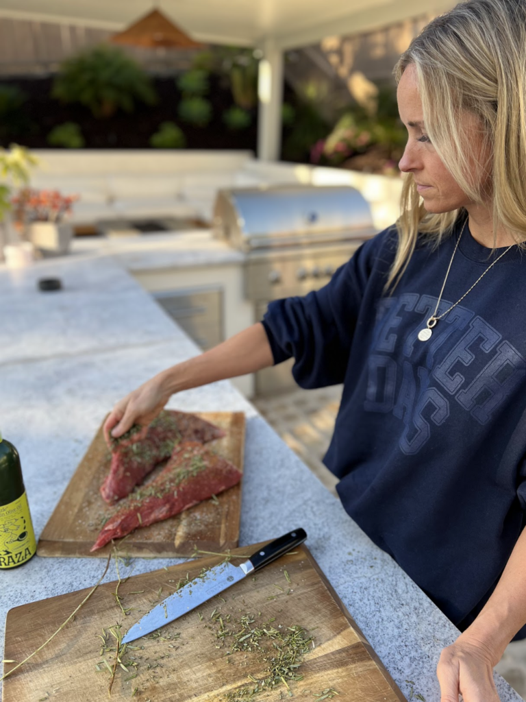 A woman seasons raw meat with herbs on a wooden cutting board outdoors. A knife and bottle of olive oil are on the counter, capturing exactly Why I Love My Outdoor Kitchen. A barbecue grill is in the background, setting the perfect scene for al fresco cooking.