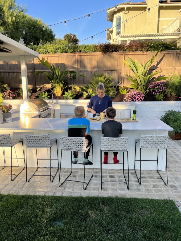 A woman prepares food at an outdoor kitchen island while two children sit on barstools watching her. Lush plants, flowers, and patio lights decorate the backyard setting, perfectly capturing why I love my outdoor kitchen.